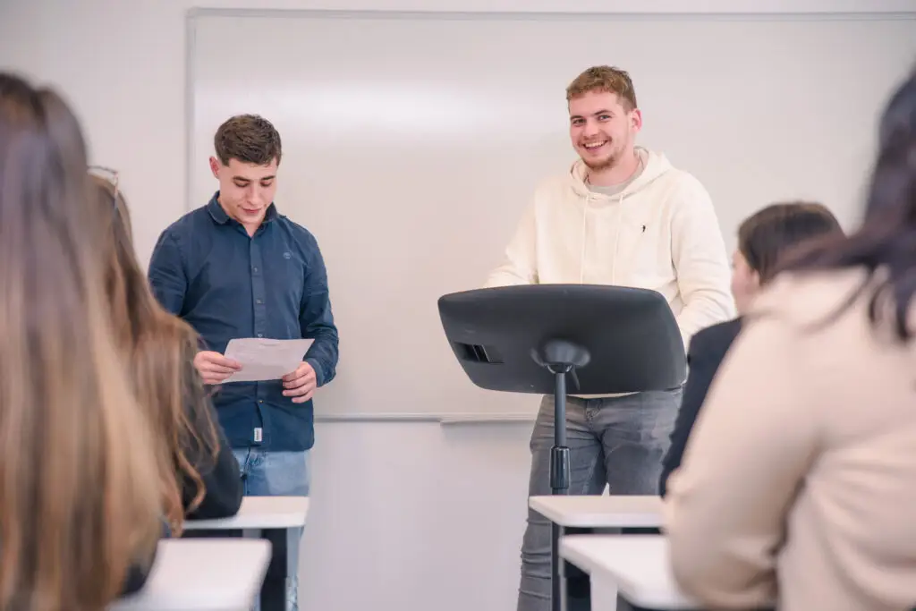 VIGNETTE – BACH RACDI Photo de deux étudiants passant un oral devant le reste de leur classe.